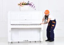 Busy worker in orange protective helmet and blue overall moving grand piano isolated on white background. White piano with flower vase on top in empty room