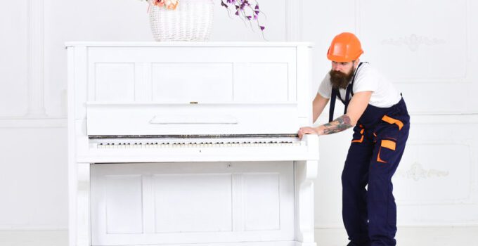 Busy worker in orange protective helmet and blue overall moving grand piano isolated on white background. White piano with flower vase on top in empty room